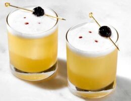 Två gula sour-cocktails med skum och körsbär på cocktailpinnar