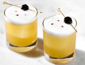 Två gula sour-cocktails med skum och körsbär på cocktailpinnar