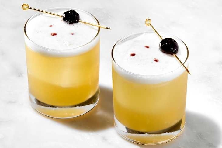 Två gula sour-cocktails med skum och körsbär på cocktailpinnar